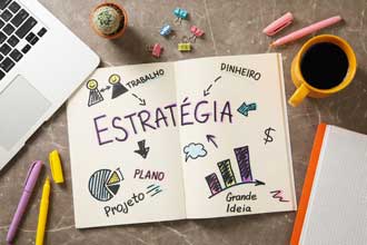 Caderno aberto com a palavra “Estratégia” escrita à mão e ilustrações sobre trabalho, dinheiro, plano e projeto, ao lado de laptop, café e materiais de escritório | Mais-R
